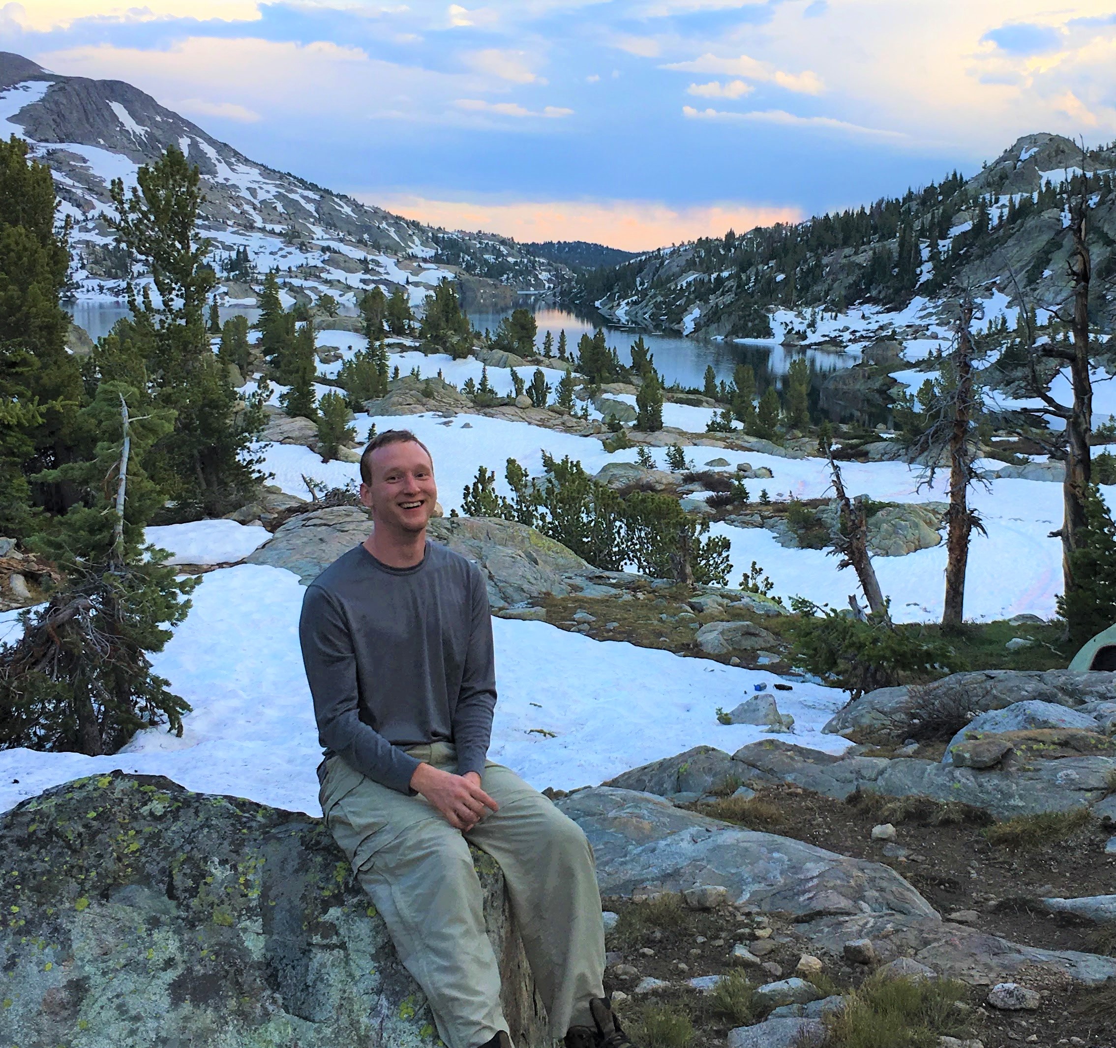 Photograph of Matthew Pintar sitting on a rock surrounded by rocks, snow, and mountains with a lake behind him.